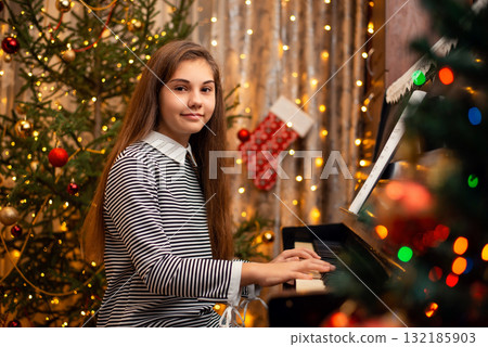 Young female pianist with long hair in a dress sitting near the piano and looking into the camera. Christmas decoration all around in the room, festive mood. 132185903