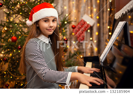 Happy girl in Christmas hat sitting near decorated fir tree and playing the piano. New Year, holidays, time at home with family concept. 132185904