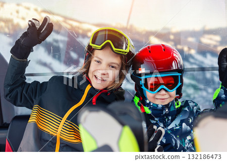 Portrait of two happy kid boys enjoy having fun sit ski lift gondola at alpine mountain skiing resort on bright winter day. Teen beginner skier rest of training in ski school 132186473