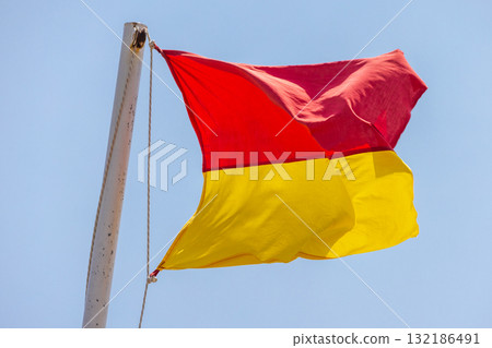 Bright red and yellow flag flutters on a white flagpole, set against a crisp blue sky Bright red and yellow flag flutters on a white flagpole, set against a crisp blue sky 132186491