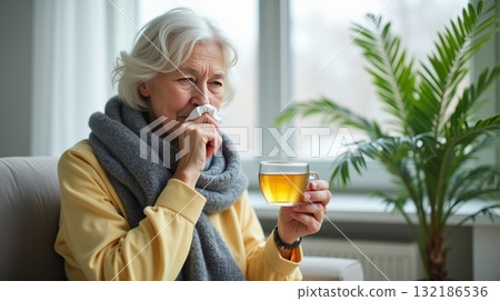 Elderly woman in scarf holding tea cup and tissue, sitting indoors with plant in background, concept of cold and flu relief. Concept of cold treatment Elderly woman in scarf holding tea cup and tissue, sitting indoors with plant in background, concept of cold and flu relief. Concept of cold treatment 132186536