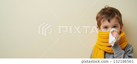 Young boy in a yellow scarf holding a tissue, set against a plain background, representing a concept of cold and flu. Common cold concept. Copy space. Young boy in a yellow scarf holding a tissue, set against a plain background, representing a concept of cold and flu. Common cold concept. Copy space. 132186561
