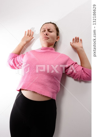 Woman in pink top and black leggings leans against white wall with eyes closed and pained expression. Woman in pink top and black leggings leans against white wall with eyes closed and pained expression. 132186629