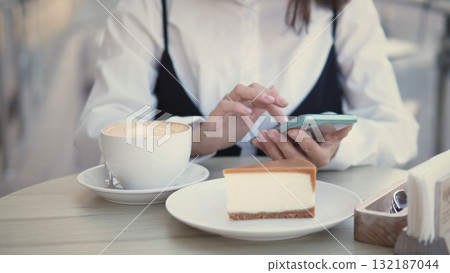 A business woman sits at a cafe table with a cup of coffee and a slice of cheesecake. She is focused on her smartphone, likely managing work tasks or messages. 132187044