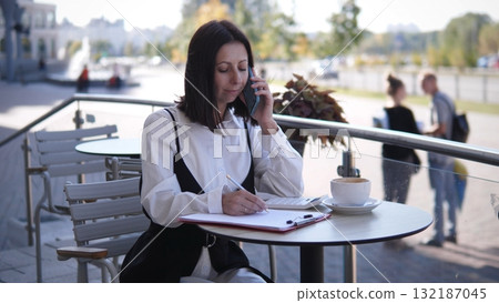 In a bright outdoor cafe setting, a business woman speaks on her phone while writing notes. She enjoys a cup of coffee as people stroll by in the background on a sunny day. 132187045