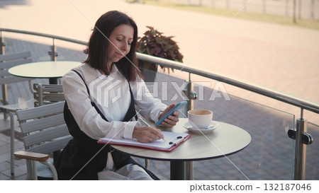 A business woman sits at an outdoor cafe table in the morning, writing notes in a notebook while occasionally checking her phone. A cup of coffee sits beside her. 132187046
