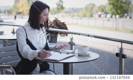 A woman sits at a table outside a cafe, focusing on her work as she takes notes. She enjoys a cup of coffee with a relaxed atmosphere around her. A woman sits at a table outside a cafe, focusing on her work as she takes notes. She enjoys a cup of coffee with a relaxed atmosphere around her. 132187048