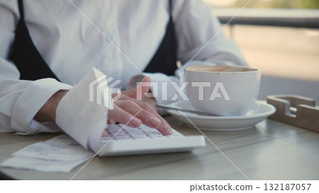 A focused business woman types on her laptop at a cafe while sipping coffee. She appears engaged in her work surrounded by a relaxed atmosphere. 132187057