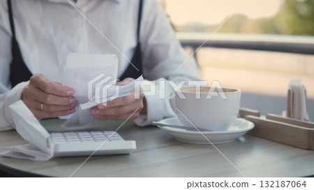 A woman dressed in a white blouse examines a receipt at a cafe table. She sits with a calculator and a cup of coffee, focused on her tasks in the pleasant afternoon sun. 132187064