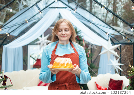 Smiling blonde woman holding homemade Christmas cake in festive kitchen setting Smiling blonde woman holding homemade Christmas cake in festive kitchen setting 132187294