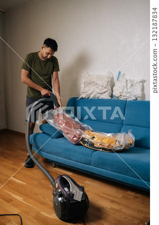 Vertical shot of man using vacuum cleaner to remove air from vacuum storage bag with clothes and placing it on sofa in living room, organizing seasonal wardrobe for winter. Concept of household chores Vertical shot of man using vacuum cleaner to remove air from vacuum storage bag with clothes and placing it on sofa in living room, organizing seasonal wardrobe for winter. Concept of household chores 132187834