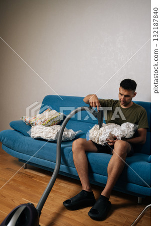Portrait of man sitting on sofa using vacuum cleaner to suck air out of storage bag containing bedding, reducing storage space and protecting from dust and moisture. Concept of household chores. 132187840