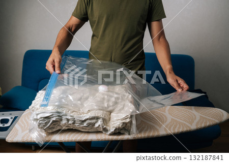 Close-up cropped shot of man closing vacuum storage bag placed on ironing board, containing bedding or out-of-season clothes, sealing for efficient space-saving storage. Concept of household chores. Close-up cropped shot of man closing vacuum storage bag placed on ironing board, containing bedding or out-of-season clothes, sealing for efficient space-saving storage. Concept of household chores. 132187841
