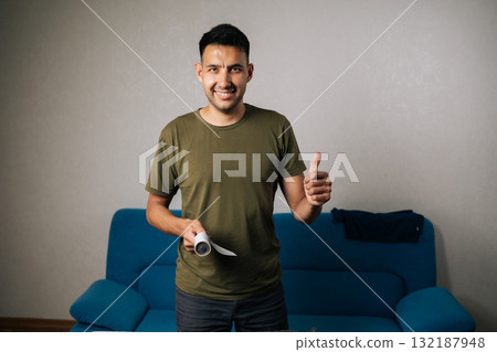 Vertical portrait of positive housekeeper male using sticky lint roller to remove dust, hair or fluff from clothes, ensuring they clean, showing thumbs up gesture, smiling looking at camera. Vertical portrait of positive housekeeper male using sticky lint roller to remove dust, hair or fluff from clothes, ensuring they clean, showing thumbs up gesture, smiling looking at camera. 132187948