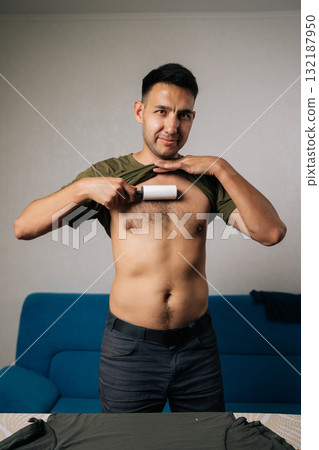 Vertical portrait of smiling young man lifting shirt and using sticky lint roller to remove hair and fluff from chest at home, showcasing humorous approach to personal grooming. 132187950