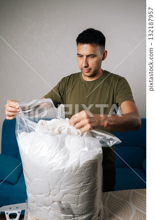 Vertical portrait of young man closing large transparent vacuum storage bag filled with white bedding, using double-zip closure to create airtight seal for optimal space efficiency and protection. 132187957