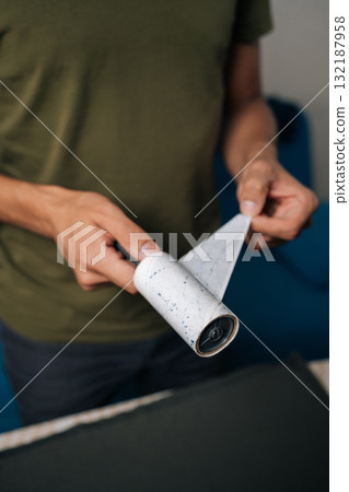 Vertical cropped shot of man peeling off used layer of sticky lint roller, preparing to clean clothes from dust, hair and fibers, ensuring garments remain tidy and fresh. Concept of household chores. Vertical cropped shot of man peeling off used layer of sticky lint roller, preparing to clean clothes from dust, hair and fibers, ensuring garments remain tidy and fresh. Concept of household chores. 132187958