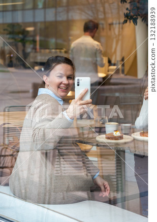 Smiling woman takes a photo with her smartphone through a cafe window, surrounded by coffee cups and pastries 132188939