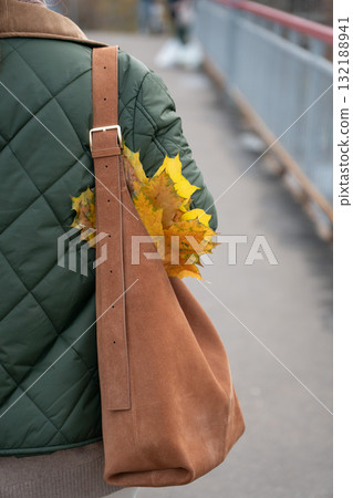 Close-up of a person wearing a green quilted jacket and brown suede bag with yellow autumn leaves tucked under the strap 132188941