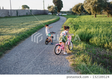two small girls riding bicycle on a small road outdoors during sunset 132189124