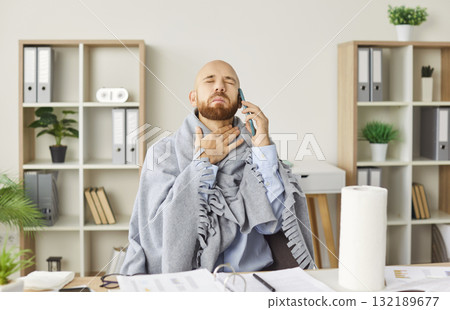Young sick man suffering from a sore throat sitting on workplace in office and talking by mobile phone. 132189677