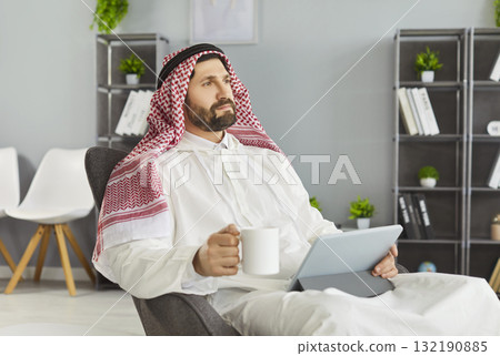 Calm executive in arab traditional dress holding coffee and tablet in casual office Calm executive in arab traditional dress holding coffee and tablet in casual office 132190885