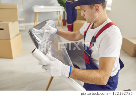 Portrait of young male mover packing furniture with plastic wrap in the living room 132192785