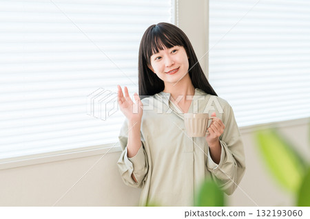 Young Asian woman greeting someone by the window with a hot drink, tea, coffee Young Asian woman greeting someone by the window with a hot drink, tea, coffee 132193060