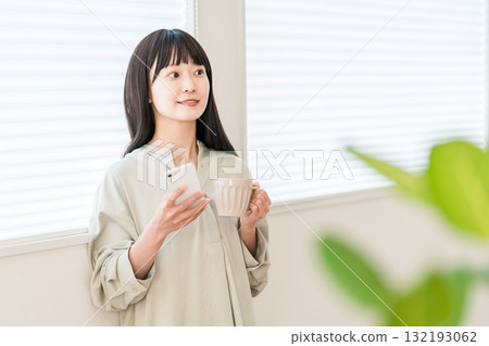 年輕的亞洲女性一邊在窗邊喝熱飲、茶或咖啡，一邊看著智慧型手機 132193062
