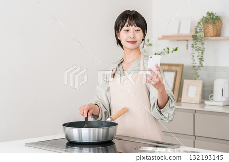 一位年輕的亞洲女子繫著圍裙在廚房做飯,一邊看著智慧型手機 一位年輕的亞洲女子繫著圍裙在廚房做飯,一邊看著智慧型手機 132193145