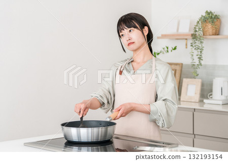 A young Asian woman in an apron cooking in front of an induction stove in the kitchen, thinking and worrying A young Asian woman in an apron cooking in front of an induction stove in the kitchen, thinking and worrying 132193154