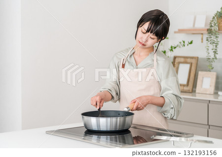 A young Asian woman in an apron cooking in front of an induction stove in the kitchen, thinking and worrying 132193156