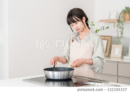 A young Asian woman in an apron cooking in front of an induction stove in the kitchen, thinking and worrying 132193157