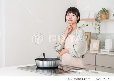 一位繫著圍裙的年輕亞洲女子在廚房的電磁爐前做飯,思考著,憂心忡忡 一位繫著圍裙的年輕亞洲女子在廚房的電磁爐前做飯,思考著,憂心忡忡 132193230