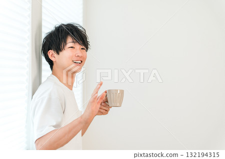 年輕的亞洲男子在窗邊用熱飲、茶或咖啡向某人打招呼 年輕的亞洲男子在窗邊用熱飲、茶或咖啡向某人打招呼 132194315