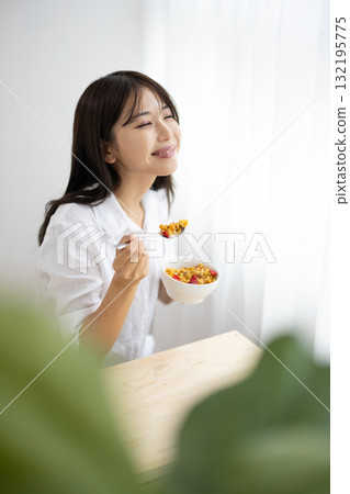 一位年輕女子在窗邊沐浴著明亮的陽光，微笑著吃著麥片 132195775