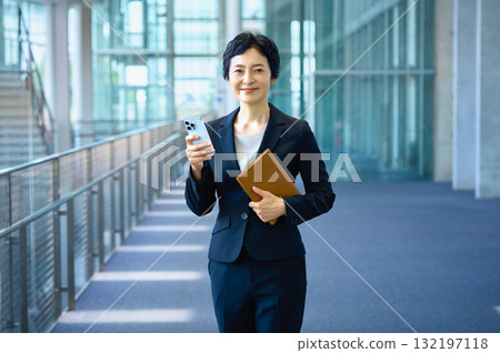 Smiling businesswoman walking through the office while looking at her smartphone (career, middle-aged, one person) Smiling businesswoman walking through the office while looking at her smartphone (career, middle-aged, one person) 132197118