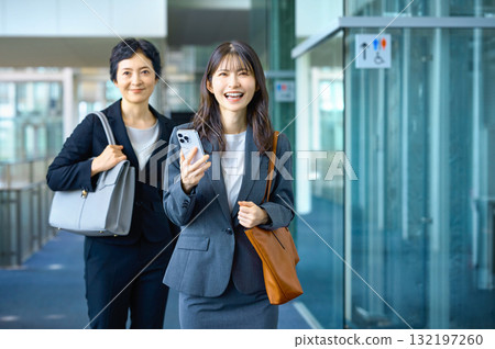 Two businesswomen walking through the office with a smile and holding a smartphone (young and middle-aged, career, two people) 132197260