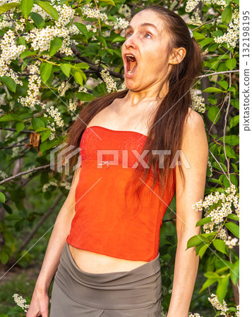 Young woman sneezes in the park against the background of a flowering tree. Allergy to pollen concept. 132198195