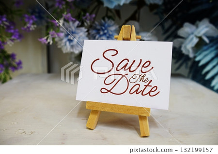 Save the Date text write on paper card with wooden easel and flowers decoration on wooden background Save the Date text write on paper card with wooden easel and flowers decoration on wooden background 132199157