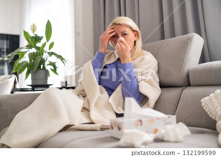 Middle aged blonde woman sitting on sofa at home, feeling unwell with cold and headache. Wrapped in blanket, resting while recovering. Middle aged blonde woman sitting on sofa at home, feeling unwell with cold and headache. Wrapped in blanket, resting while recovering. 132199599