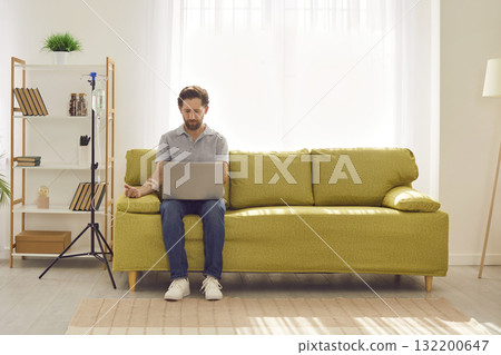 Young man working with laptop at home while receiving IV drip infusion and vitamin therapy 132200647