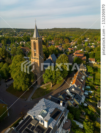 Aerial view of town with central Gothic style church, pointed steeple, red and gray rooftops, tree lined streets, and parked cars blending history with modern life. 132200970