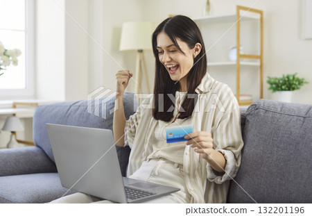 Young happy woman purchasing online seated on sofa with laptop and card 132201196