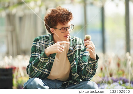 Sitting and holding the little cactus. Young man with curly hair and in glasses is in greenhouse 132202960
