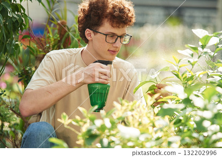 With green watering bottle. Young man with curly hair and in glasses is in greenhouse With green watering bottle. Young man with curly hair and in glasses is in greenhouse 132202996
