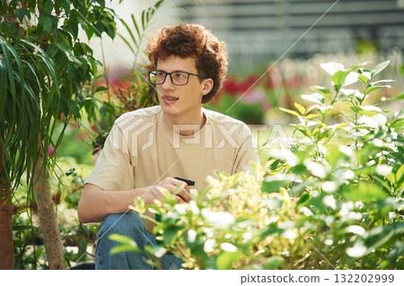 Growing flowers. Conception of watering. Young man with curly hair and in glasses is in greenhouse 132202999