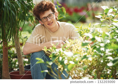 Growing flowers. Conception of watering. Young man with curly hair and in glasses is in greenhouse 132203000