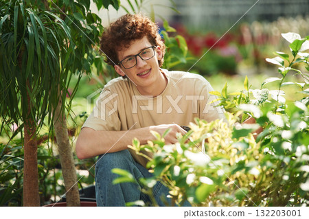 Growing flowers. Conception of watering. Young man with curly hair and in glasses is in greenhouse Growing flowers. Conception of watering. Young man with curly hair and in glasses is in greenhouse 132203001