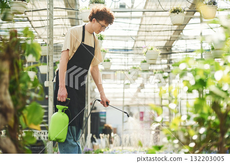 Watering the plants. Young man with curly hair and in glasses is in greenhouse 132203005